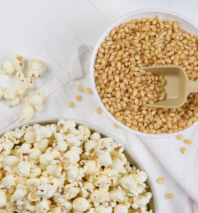 A white bowl of popcorn kernels next to a bowl of stovetop popcorn on a white tablecloth.